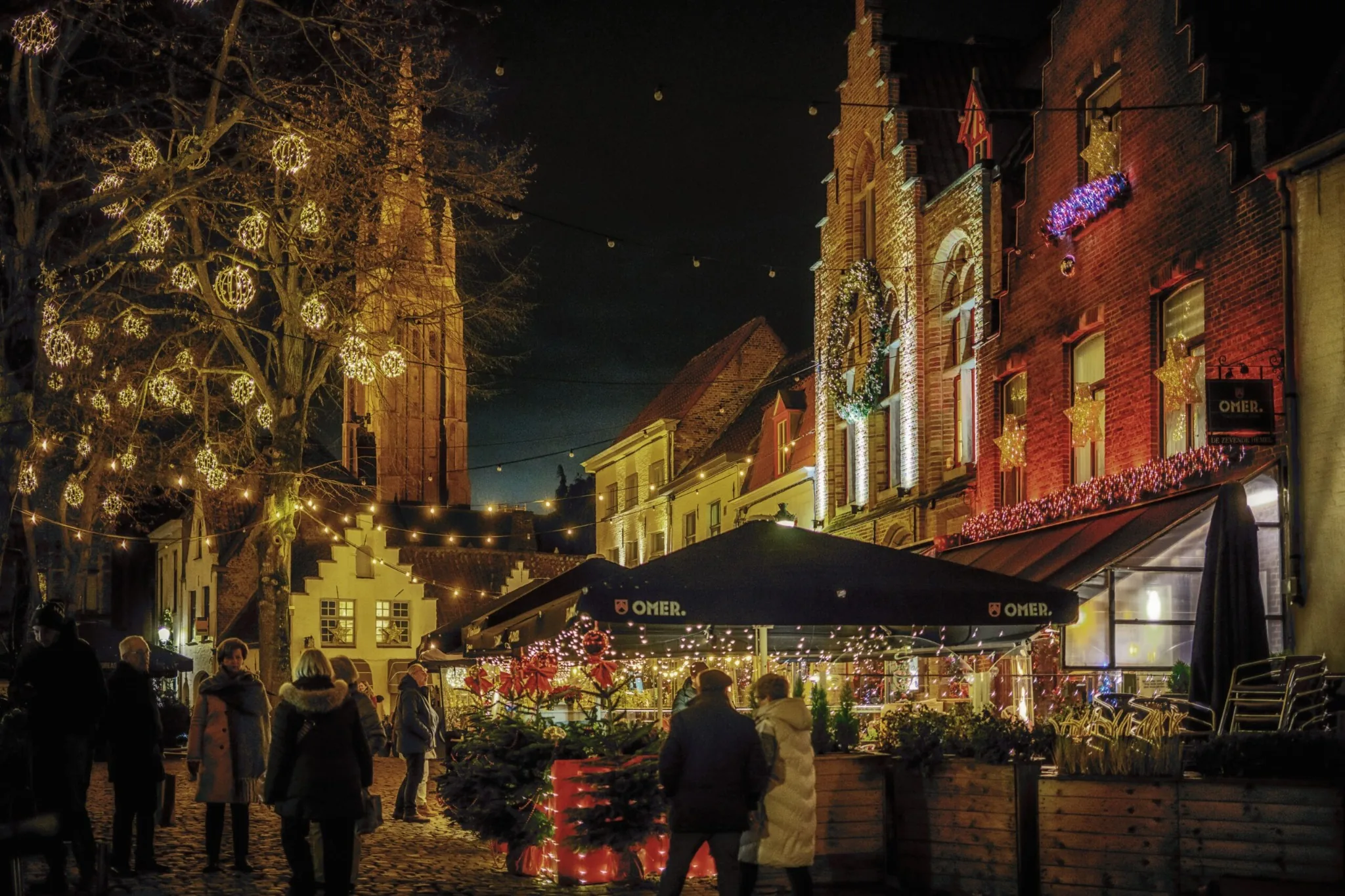 Festliche Abendstraße in Brügge mit Lichtern in den Bäumen, einer Terrasse und einem Kirchturm